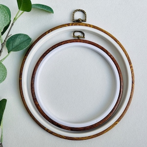 以下が含まれることがあります： 2つの刺繍枠が重なっています。外側の枠は茶色で、金色の金属製のリングが付いています。内側の枠は白です。両方の枠は白い布の背景の上にあります。