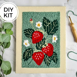 以下が含まれることがあります： パンチニードルストロベリーデザインのDIYキット。このアートワークは、白い種、緑の葉、白い花が付いた3つの赤いイチゴを、テクスチャ加工された緑色の背景に示しています。「DIY KIT」という言葉が白い円の中にあります。