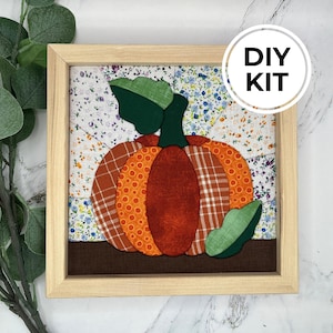 May include: A square framed fabric art piece featuring a pumpkin made of orange, red, and plaid fabric patches with green fabric leaves. The background consists of floral patterned fabric squares. A circular white label reads DIY KIT.