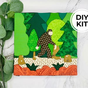 Puede incluir: Un kit de manualidades de tela que muestra una figura de Bigfoot con lunares marrones y blancos caminando por un bosque de árboles verdes. La escena incluye un suelo beige con estrellas y una base naranja con motivos florales. El texto dice DIY KIT.