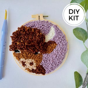 以下が含まれることがあります： DIYパンチニードルキット。茶色と白の牛が紫色の背景に描かれています。キットには、木製刺繍枠、糸、パンチニードルが含まれています。