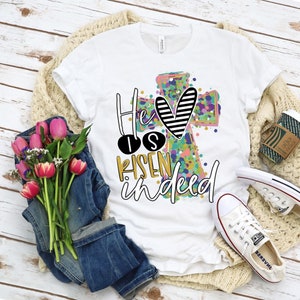 May include: White t-shirt with the text "He is Risen Indeed" in a colorful design featuring a cross and heart. The shirt is paired with blue jeans, pink tulips, and white sneakers, creating a casual, spring-themed aesthetic.