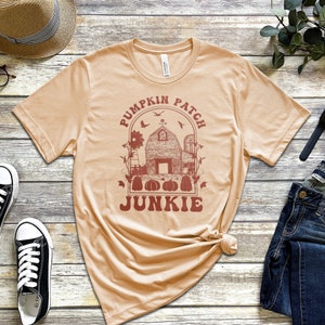 May include: A tan t-shirt with a red graphic of a pumpkin patch with a barn, pumpkins, and the text "Pumpkin Patch Junkie".