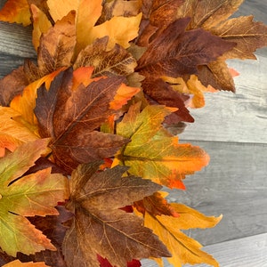May include: A close-up of a fall wreath made of artificial maple leaves in shades of orange, brown, and yellow. The leaves are arranged in a circular pattern and are overlapping.