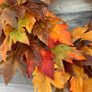 May include: A fall-themed wreath with a variety of artificial maple leaves in shades of red, orange, yellow, and brown. The leaves are arranged in a circular pattern, creating a festive and autumnal display.
