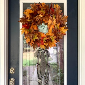 May include: A fall-themed wreath made of artificial leaves in shades of orange, red, and brown hangs on a blue front door with a glass panel.