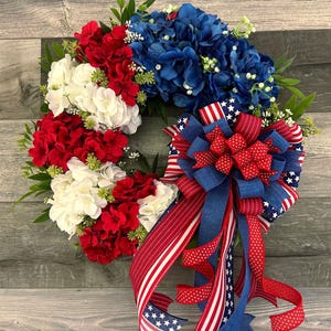 May include: A patriotic wreath featuring red, white, and blue hydrangeas and a large bow. The bow is made of ribbons with stars and stripes patterns. Green foliage accents the floral arrangement, creating a festive and decorative piece.