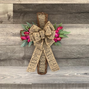 May include: A burlap cross with a floral wreath and a burlap bow. The cross has the words "God so loved the world that He gave His only begotten Son, that whoever believes in Him" written on it.