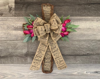 Op de afbeelding: Een jute kruis met een bloemenkrans en een jute strik. Op het kruis staat de tekst "God heeft de wereld zo liefgehad, dat Hij Zijn enige Zoon gegeven heeft, opdat een ieder die in Hem gelooft, niet verloren gaat, maar eeuwig leven heeft." geschreven.