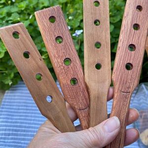 May include: Four wooden paddles with holes drilled in them. The paddles are brown with a reddish hue and are held in a hand.