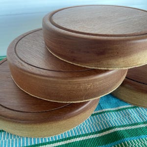 May include: Four wooden coasters with a dark brown finish are stacked. Each coaster has a circular design with a raised edge and a lighter base. The coasters are arranged on a blue and green striped surface.