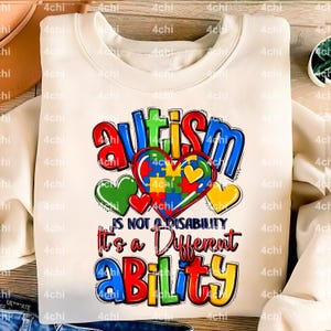 May include: A white sweatshirt with a colorful graphic design that says "autism is not a disability it's a different ability". The graphic includes a heart made of puzzle pieces and smaller hearts in different colors.