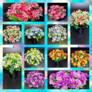 May include: A selection of artificial flower arrangements in various colours, including pink, red, orange, yellow, white, and purple. Each arrangement is in a black pot, with green foliage. The flowers are daisy-like in appearance.