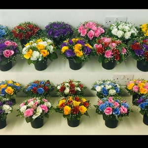 May include: A collection of artificial rose arrangements in small black pots. The roses are in various colours, including blue, red, yellow, pink, purple, and white, with small white accent flowers.