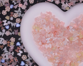 May include: A heart-shaped dish filled with pink, white, and yellow glitter and butterfly-shaped charms. The dish is on a black background with more glitter and charms.