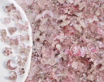 May include: A large assortment of pink glitter resin charms in various shapes, including butterflies, flowers, hearts, and moons. The charms are scattered on a white surface.