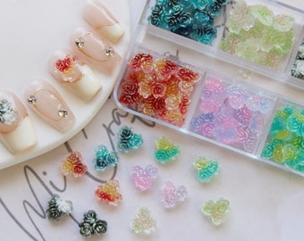 May include: Assortment of colorful flower-shaped nail charms in a variety of colors, including red, pink, green, blue, and white. The charms are arranged in a clear plastic container with multiple compartments.