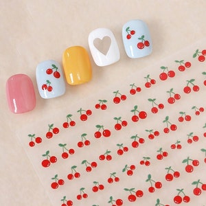 May include: A sheet of nail art stickers with a repeating pattern of red cherries with green stems and leaves. There are five painted fingernails with cherry designs on a light background.