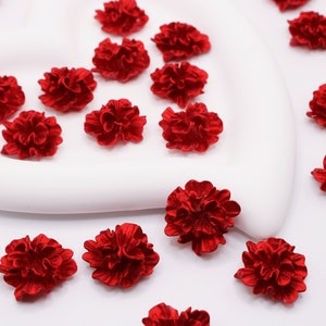 May include: A collection of small, red, fabric carnation flowers. The flowers are arranged on a white surface.