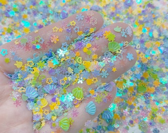 May include: A variety of iridescent glitter shapes in pastel colors, including stars, snowflakes, seashells, and hearts. The glitter is scattered on a hand, creating a colorful and sparkly display.