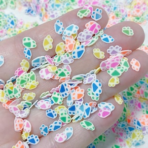 May include: A variety of colorful paw print shaped sprinkles. The sprinkles are made of a white base with different colored accents. The colors include blue, green, yellow, orange, pink, and purple.