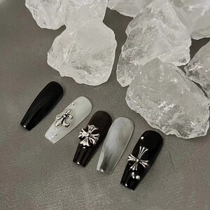 May include: A set of five long, coffin-shaped artificial nails. The nails are black, white, and gray, with silver cross charms and small rhinestones. The nails are arranged on a gray surface.