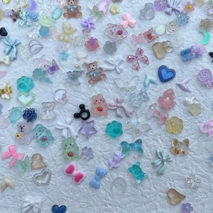 May include: Assortment of colorful resin charms in various shapes, including animals, hearts, stars, butterflies, and bows. The charms are scattered on a white background.
