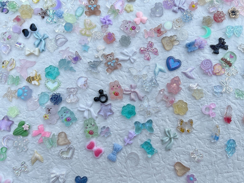 May include: Assortment of colorful resin charms in various shapes, including animals, hearts, stars, butterflies, and bows. The charms are scattered on a white background.