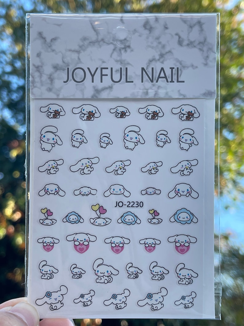 Op de afbeelding: Een vel met witte nagelstickers met verschillende schattige cartoon-hondendesigns. De stickers tonen honden met verschillende gezichtsuitdrukkingen, die harten, ballonnen en bloemen vasthouden. Het vel is gelabeld met "JOYFUL NAIL" en "JO-2230".