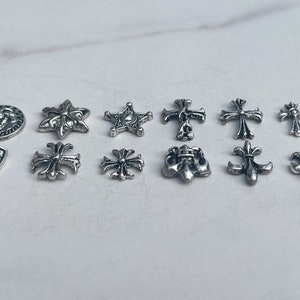 May include: A collection of silver-colored metal charms in various shapes, including crosses, stars, and fleur-de-lis designs. The charms are arranged on a light-colored surface, showcasing their intricate details and reflective surfaces. These charms are ideal for jewelry making.