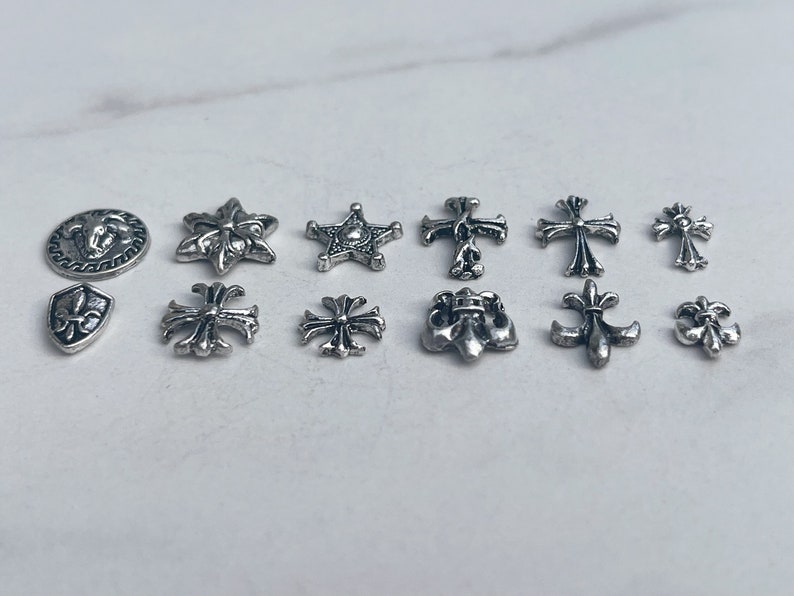 May include: A collection of silver-colored metal charms in various shapes, including crosses, stars, and fleur-de-lis designs. The charms are arranged on a light-colored surface, showcasing their intricate details and reflective surfaces. These charms are ideal for jewelry making.
