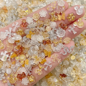 May include: Assortment of small, clear resin charms in various shapes, including crosses, flowers, butterflies, and cartoon characters. The charms are scattered on a white surface and are mostly gold and silver in color.
