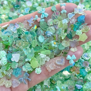 May include: A variety of green and clear resin embellishments, including flowers, butterflies, hearts, and shamrocks. The embellishments are scattered on a hand, creating a colorful and whimsical display.