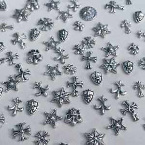 May include: A collection of small, silver-colored metal charms in various shapes, including crosses, stars, fleur-de-lis, and shields. The charms are scattered across a white surface, creating a decorative and detailed display.