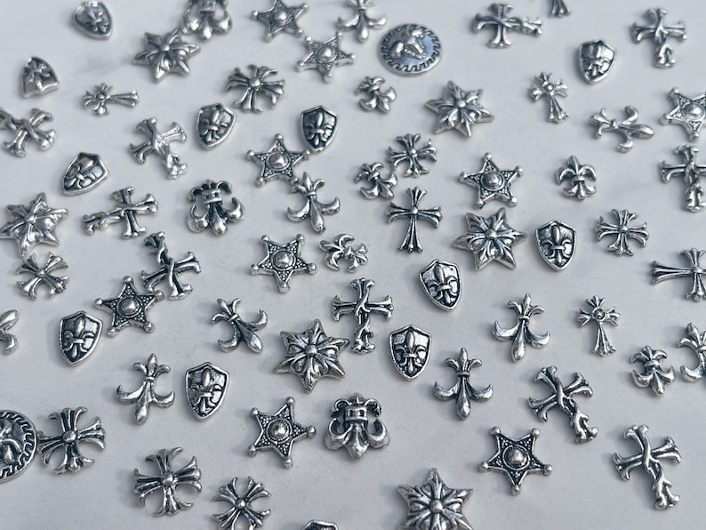 May include: A collection of small, silver-colored metal charms in various shapes, including crosses, stars, fleur-de-lis, and shields. The charms are scattered across a white surface, creating a decorative and detailed display.