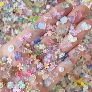 May include: Assortment of colorful, translucent resin charms in various shapes, including hearts, butterflies, stars, flowers, and bows. The charms are scattered on a hand, showcasing their iridescent and glittery details.