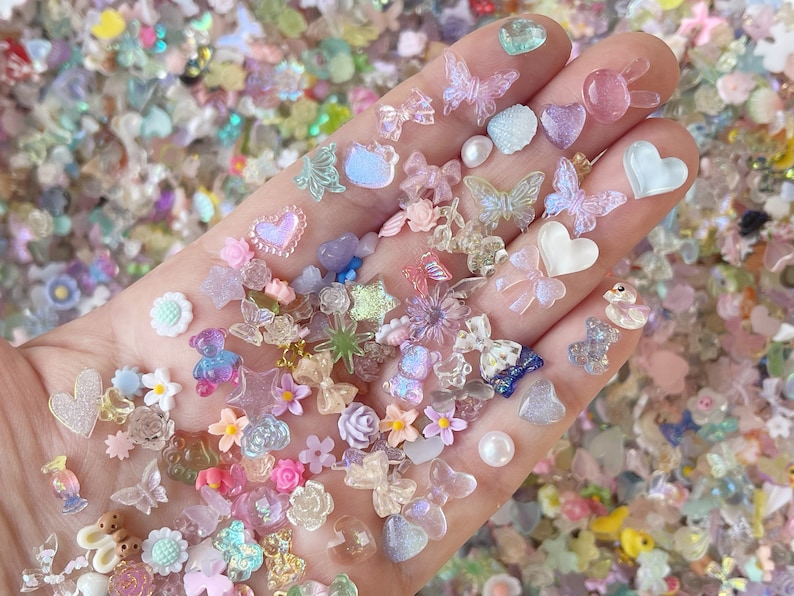 May include: Assortment of colorful, translucent resin charms in various shapes, including hearts, butterflies, stars, flowers, and bows. The charms are scattered on a hand, showcasing their iridescent and glittery details.