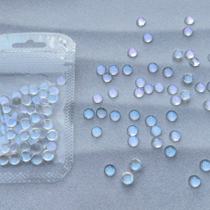 Puede incluir: Una bolsa de pl&aacute;stico transparente llena de cuentas redondas de vidrio blanco iridiscente. La bolsa est&aacute; abierta y las cuentas se est&aacute;n derramando sobre una superficie gris claro.