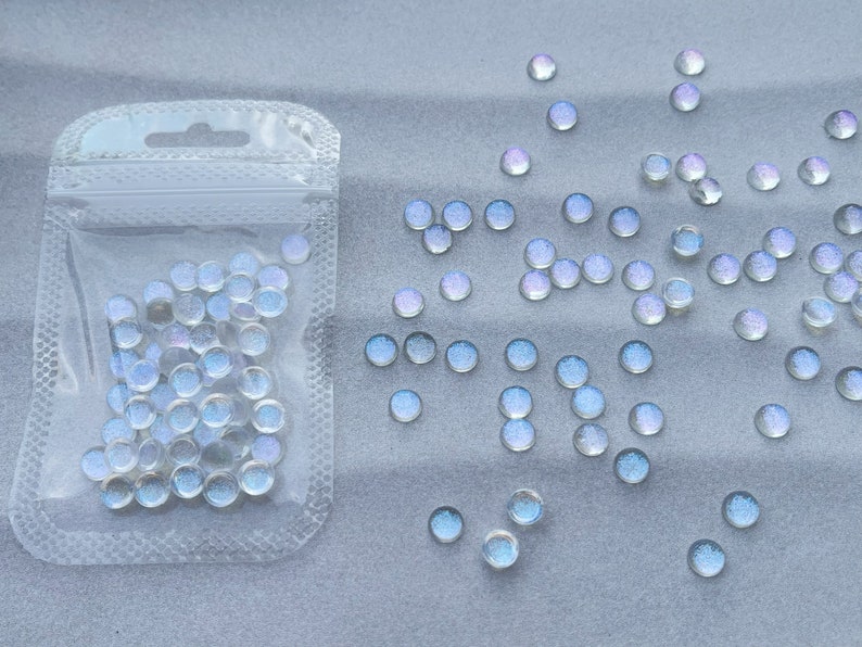 Puede incluir: Una bolsa de pl&aacute;stico transparente llena de cuentas redondas de vidrio blanco iridiscente. La bolsa est&aacute; abierta y las cuentas se est&aacute;n derramando sobre una superficie gris claro.