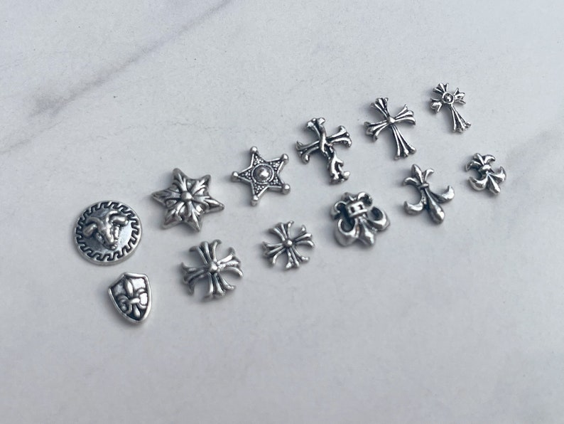 May include: A collection of small, silver-toned metal charms and embellishments. The assortment includes crosses, stars, fleur-de-lis, and round designs with intricate details. These decorative elements are ideal for jewelry making or craft projects.
