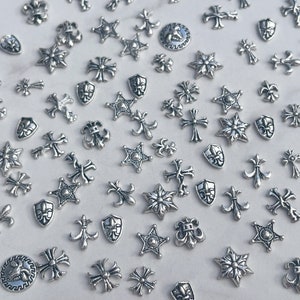May include: A collection of small, silver-colored metal charms in various shapes, including crosses, stars, fleur-de-lis, and shield designs. The charms are scattered across a white surface, creating a decorative and detailed pattern.