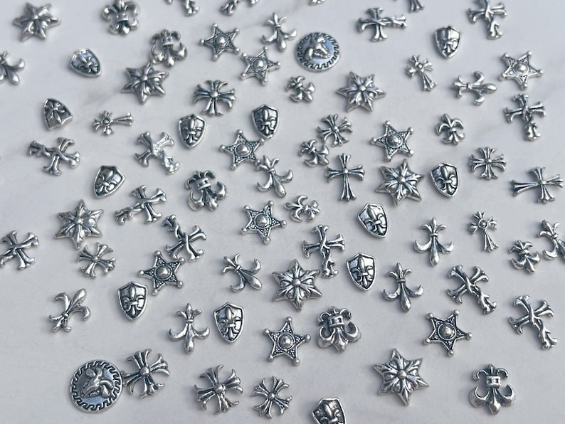 May include: A collection of small, silver-colored metal charms in various shapes, including crosses, stars, fleur-de-lis, and shield designs. The charms are scattered across a white surface, creating a decorative and detailed pattern.