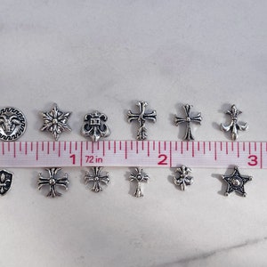 May include: A collection of small, silver-colored charms in various shapes, including crosses, stars, and fleur-de-lis. A measuring tape shows the charms are approximately 0.5 to 1 inch in size. Ideal for jewelry making or crafting.