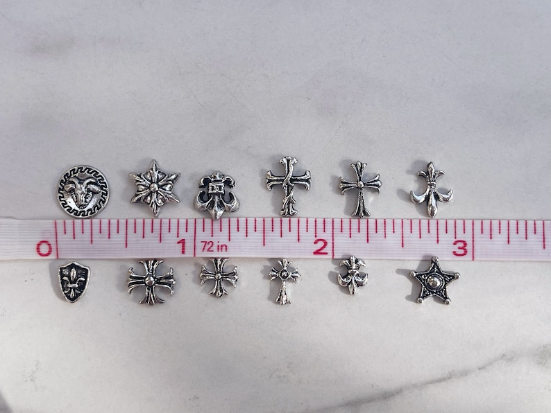 May include: A collection of small, silver-colored charms in various shapes, including crosses, stars, and fleur-de-lis. A measuring tape shows the charms are approximately 0.5 to 1 inch in size. Ideal for jewelry making or crafting.