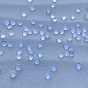 Puede incluir: Una colecci&oacute;n de cuentas de vidrio iridiscente esparcidas sobre una superficie de tela azul claro. Las cuentas son redondas y tienen un brillo sutil.