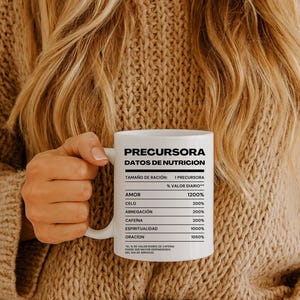 Puede incluir: Una taza de cerámica blanca con el texto "PRECURSORA DATOS DE NUTRICION" y una etiqueta de información nutricional. La etiqueta enumera los valores diarios de amor, celo, abnegación, cafeína, espiritualidad y oración. La taza es sostenida por una persona con un suéter beige.