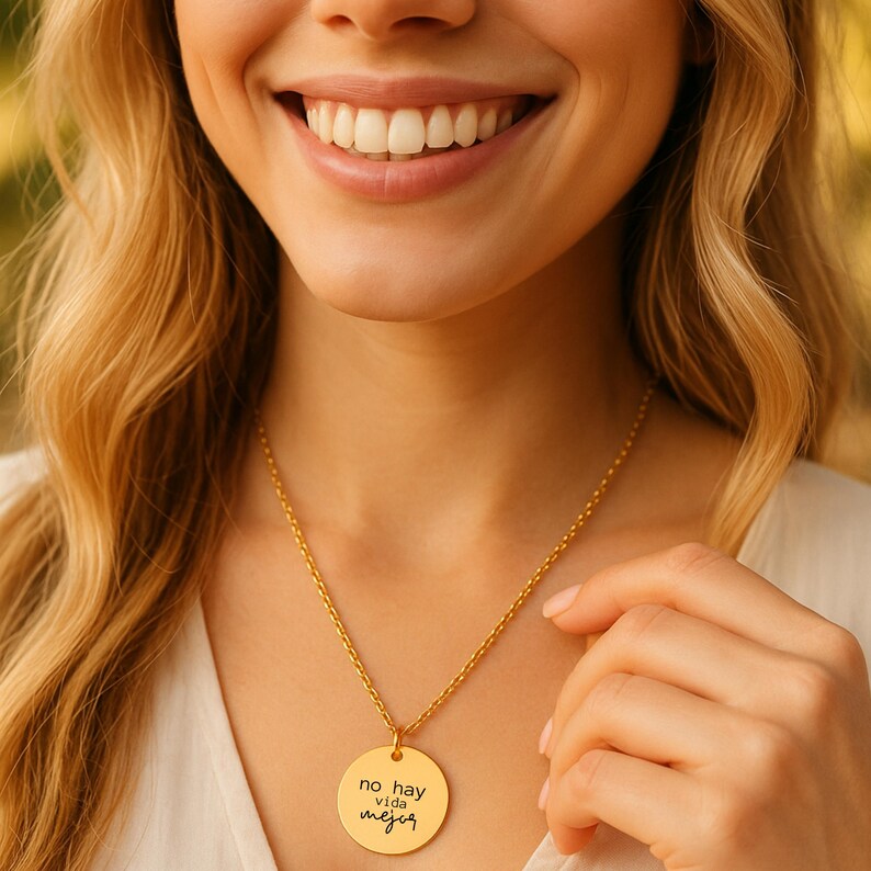 Puede incluir: Un collar de tono dorado con un colgante circular que presenta el texto grabado 'no hay vida mejor' en una fuente cursiva negra. El colgante cuelga de una delicada cadena dorada, mostrando un acabado met&aacute;lico pulido y brillante.