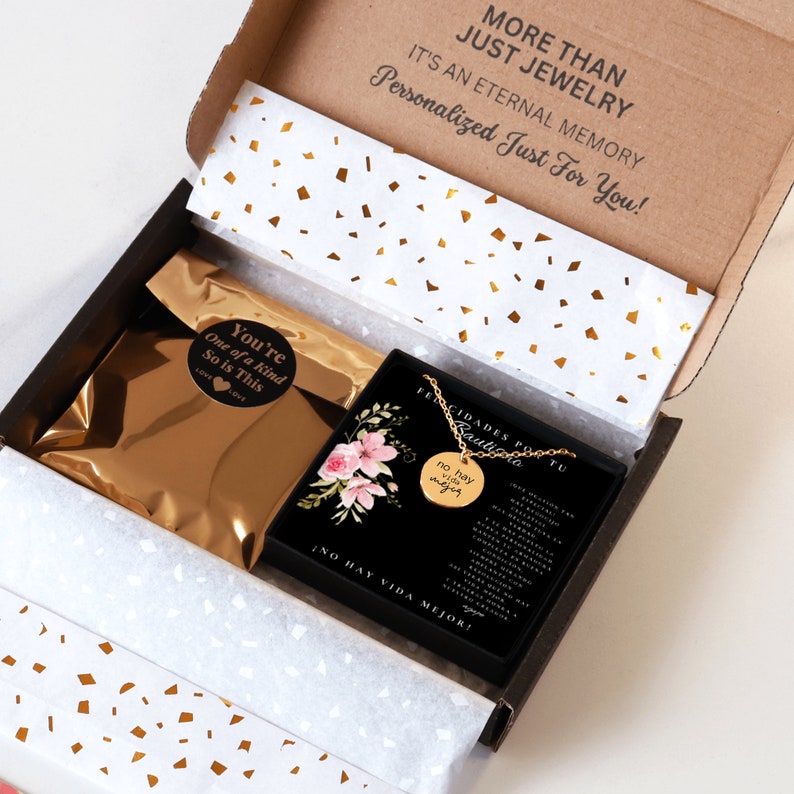 Puede incluir: Un collar con colgante circular dorado sobre una tarjeta negra con ilustraciones florales rosas y texto en espa&ntilde;ol. El collar viene en una caja de regalo de cart&oacute;n con papel de seda blanco con confeti dorado y una bolsa dorada metalizada.