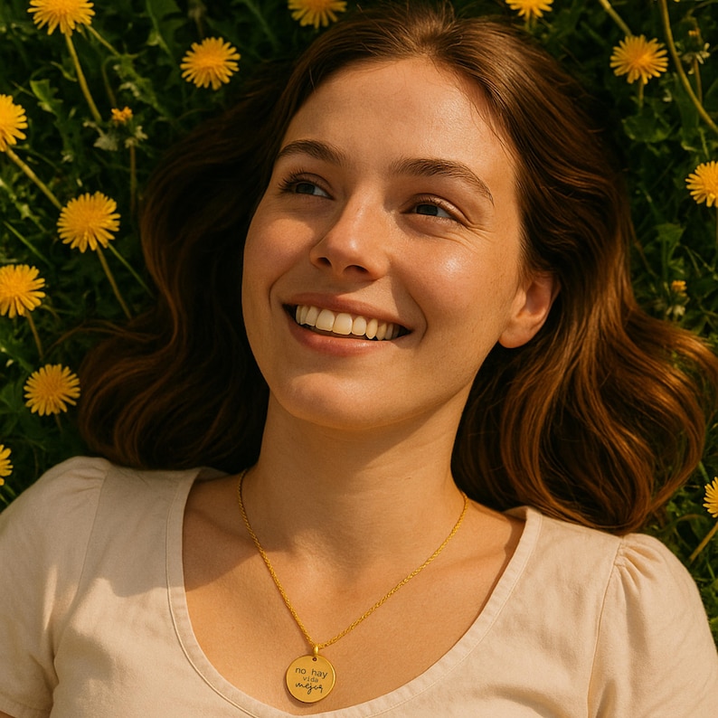 Puede incluir: Un collar con colgante circular dorado que presenta una cadena fina y un dije redondo grabado con el texto en espa&ntilde;ol 'no hay vida mejor' en letra cursiva, usado sobre una prenda clara con un fondo de flores amarillas.