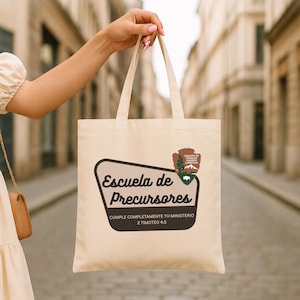 May include: Cream-colored tote bag with the text "Escuela de Precursores" in a black, rounded font. The bag also features a brown and green logo with text and a mountain and tree design. The bag is held up by a person.
